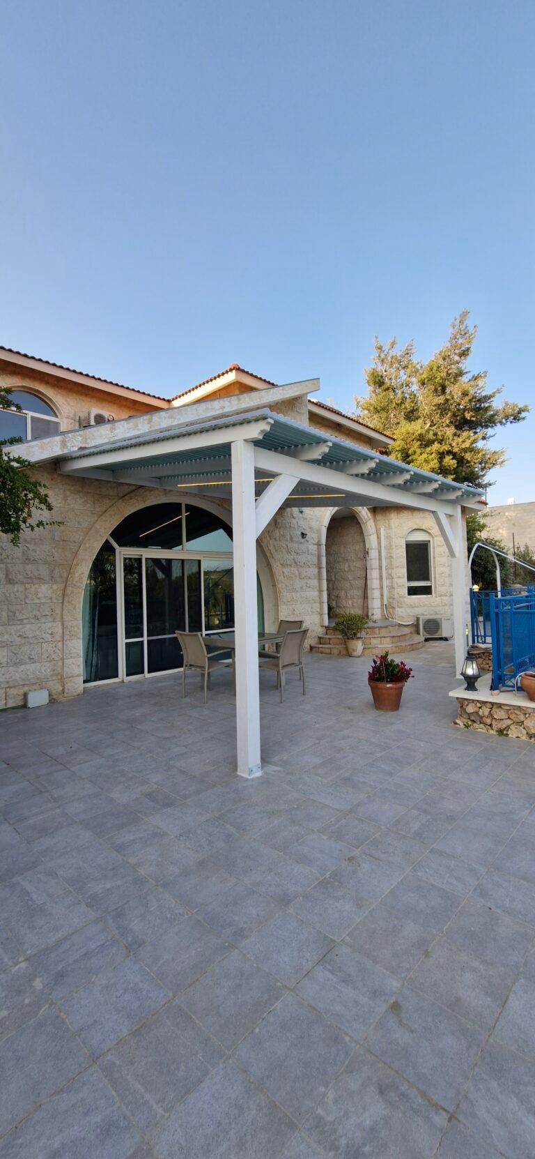 פרגולה לבנה מעל מרפסת מרוצפת בכניסה לבית אבן