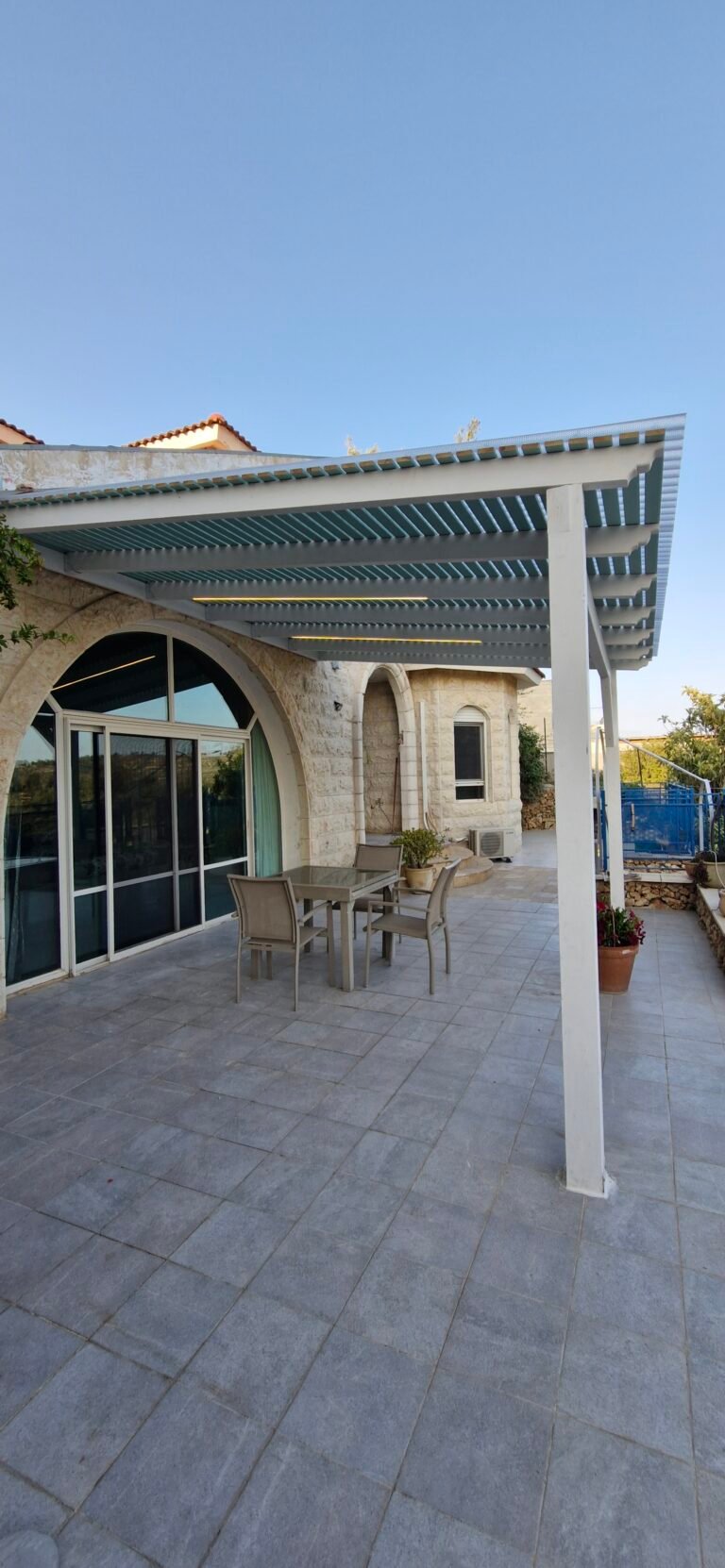 פרגולה לבנה מעל מרפסת מרוצפת ליד דלתות הזזה בבית אבן