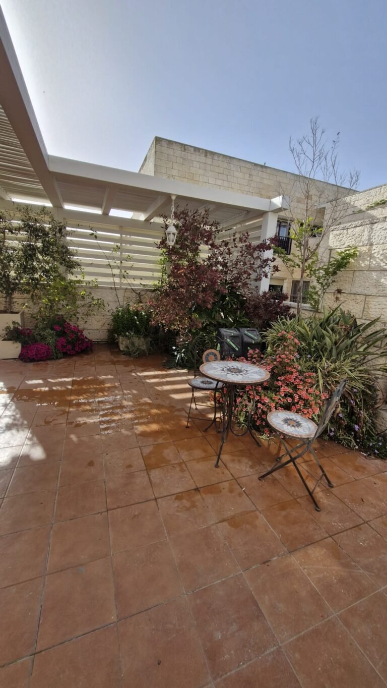 פרגולה לבנה מעל מרפסת מרוצפת עם צמחים ושולחן קטן